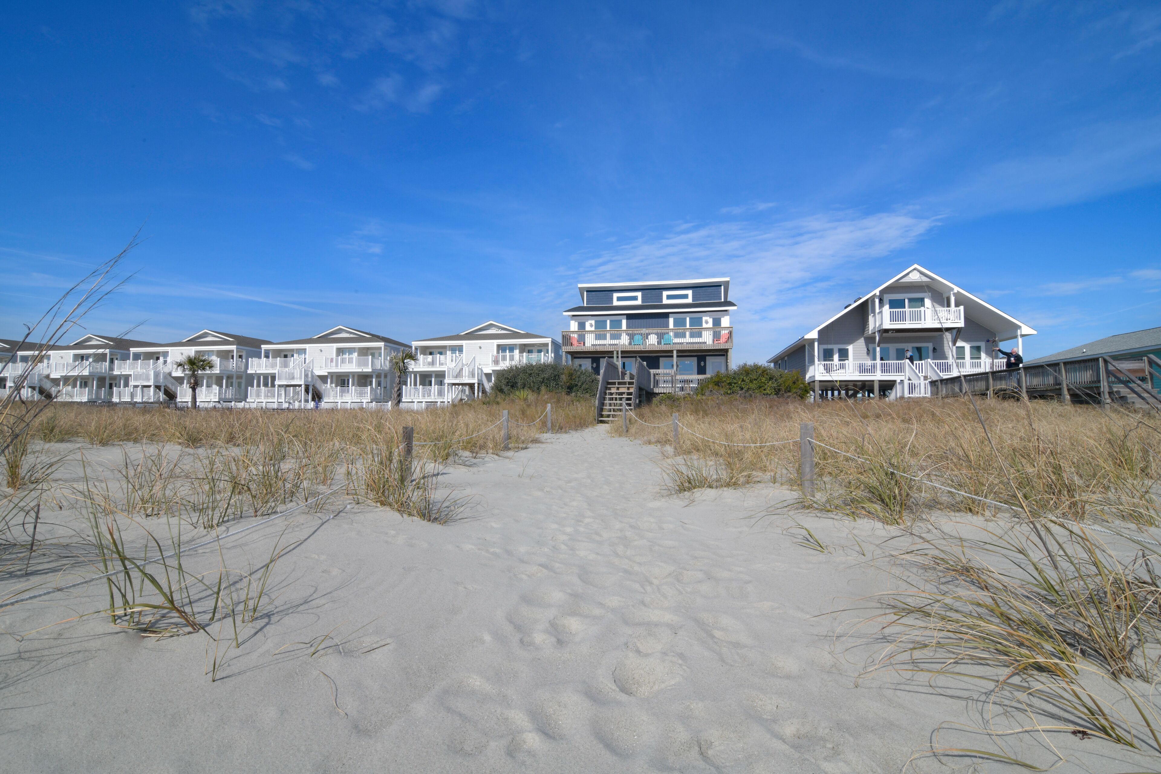 A beachfront hotel with multiple buildings, a sandy pathway leading to the entrance, and a clear blue sky.