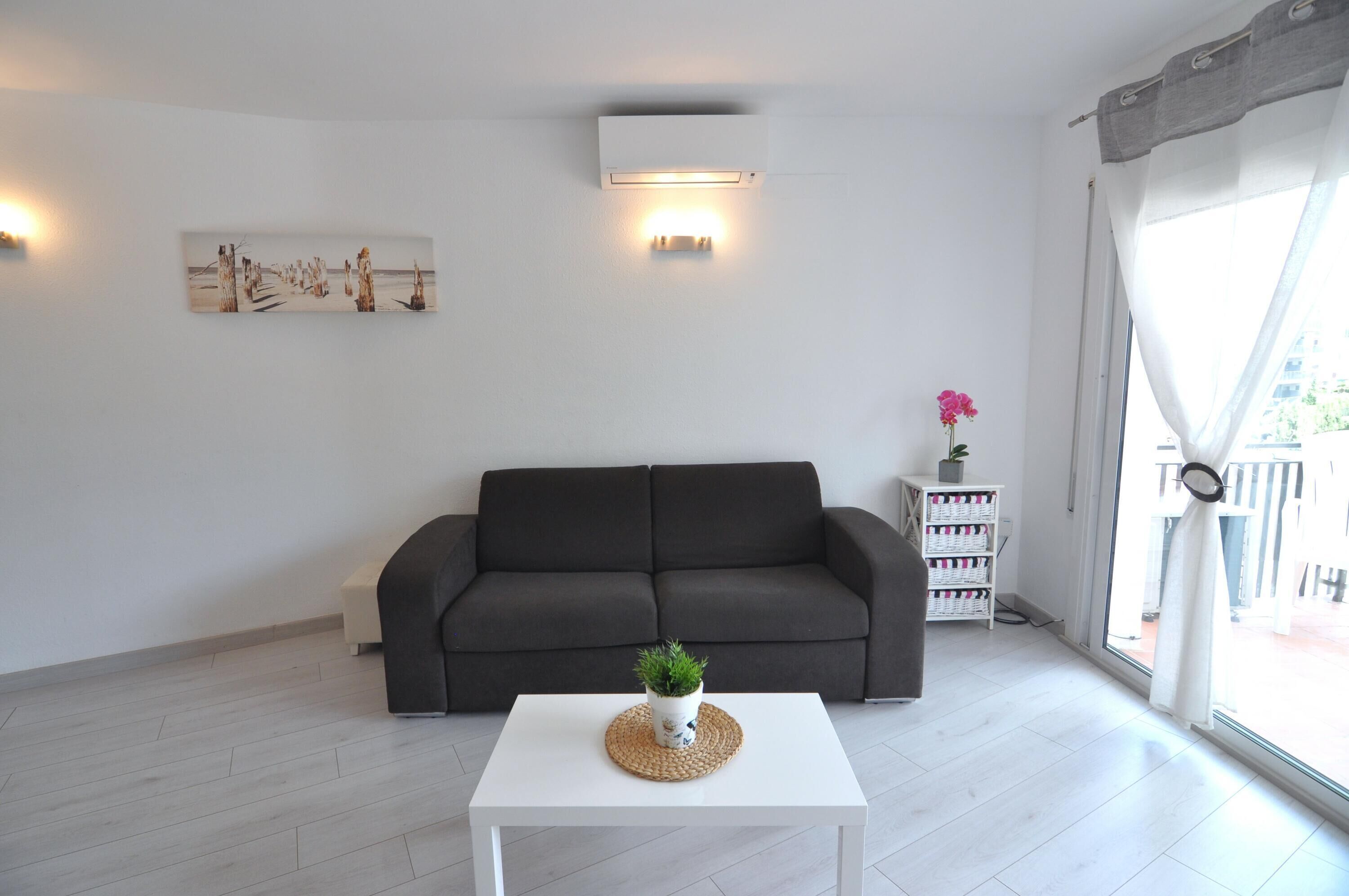 A modern living room with a dark grey sofa, a white coffee table with a plant, a wall-mounted air conditioner, and a large window with curtains.