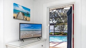 Una habitación con una televisión sobre un tocador blanco, un cuadro de playa en la pared y vistas a una piscina a través de una puerta abierta.
