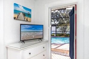 Una stanza con una TV su una cassettiera bianca, un quadro raffigurante una spiaggia al muro e vista su una piscina attraverso una porta aperta.