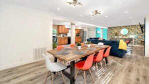Un comedor moderno con una mesa de madera y sillas coloridas, una cocina al fondo y una sala de estar con un sofá azul y un papel tapiz estampado.
