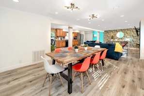 Zona pranzo moderna con tavolo di legno e sedie colorate, una cucina sullo sfondo e un salotto con divano blu e parete decorata.