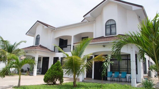 A two-story house with a white exterior, multiple windows, and a covered patio area with blue chairs. - Luxuriously Finished 4-bed House in Tema Comm 25 (Tema)