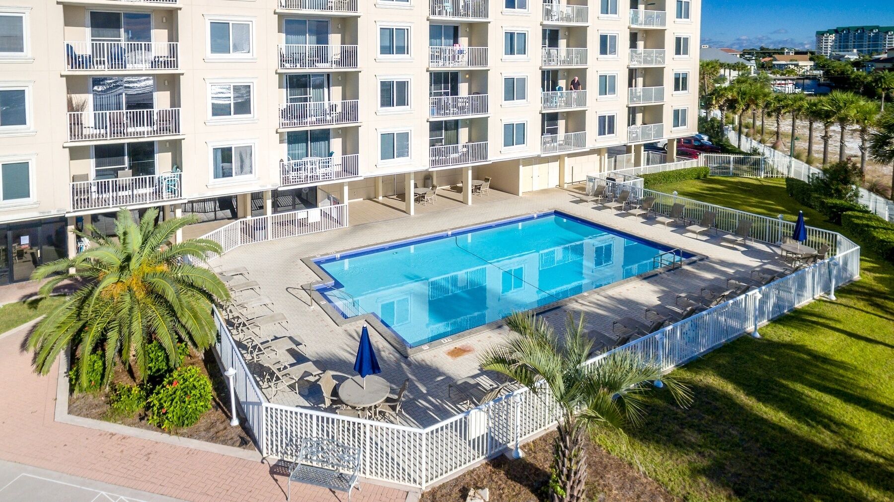 A multi-story hotel with a rectangular pool surrounded by a white fence, palm trees, and outdoor seating.