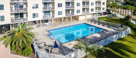 A multi-story hotel with a rectangular pool surrounded by a white fence, palm trees, and outdoor seating.