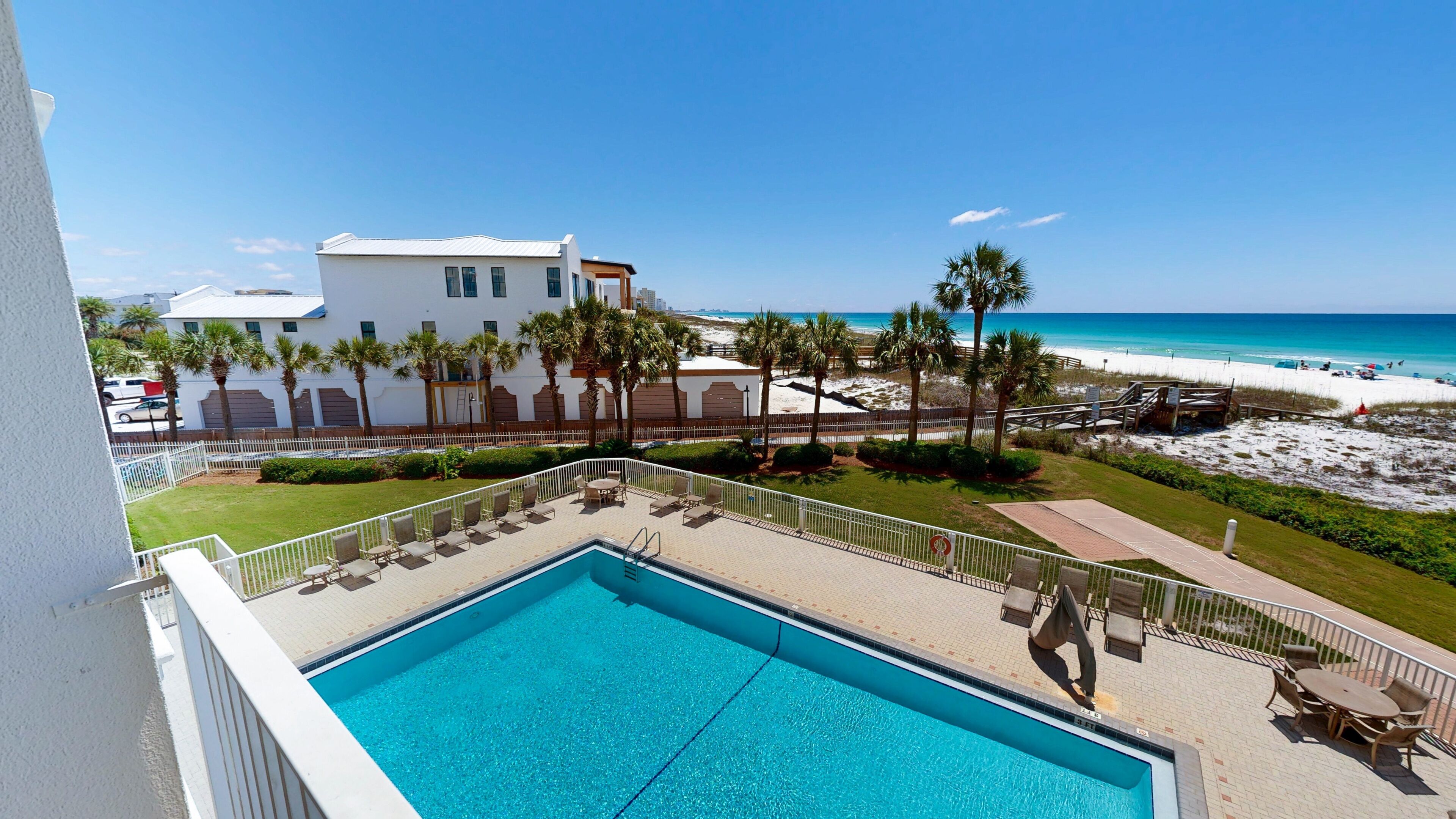 A pool area with a view of the beach, palm trees, and a white building.