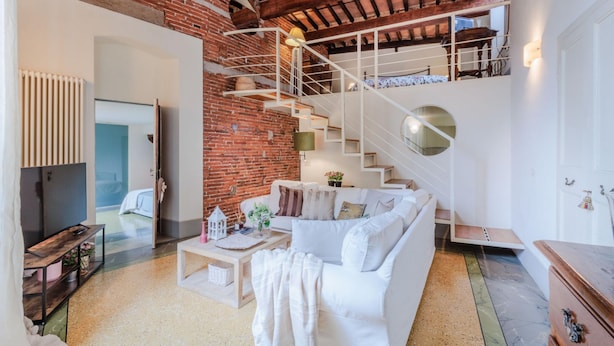 A modern living room with a white sofa, a coffee table, and a brick wall. There is a staircase leading to an upper level. - Casta Dama Apartment (Lucca)