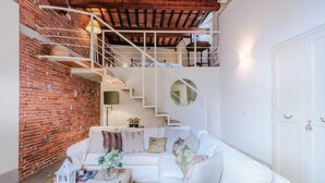 A modern living room with a white sofa, a coffee table, and a brick wall. There is a staircase with a white railing leading to an upper level. - Casta Dama Apartment (Lucca)