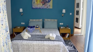 A bedroom with a blue wall, a bed with a grey quilt, two bedside tables with lamps, and a painting above the bed. - Chrismma 1-bed Apt With Private Yard & Parking (Archanes-Asterousia)