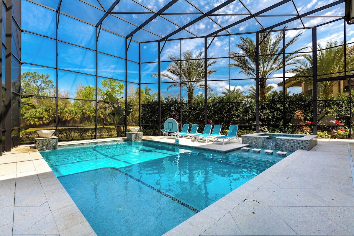 An indoor swimming pool with clear blue water, surrounded by a glass enclosure, and featuring lounge chairs and a hot tub area.
