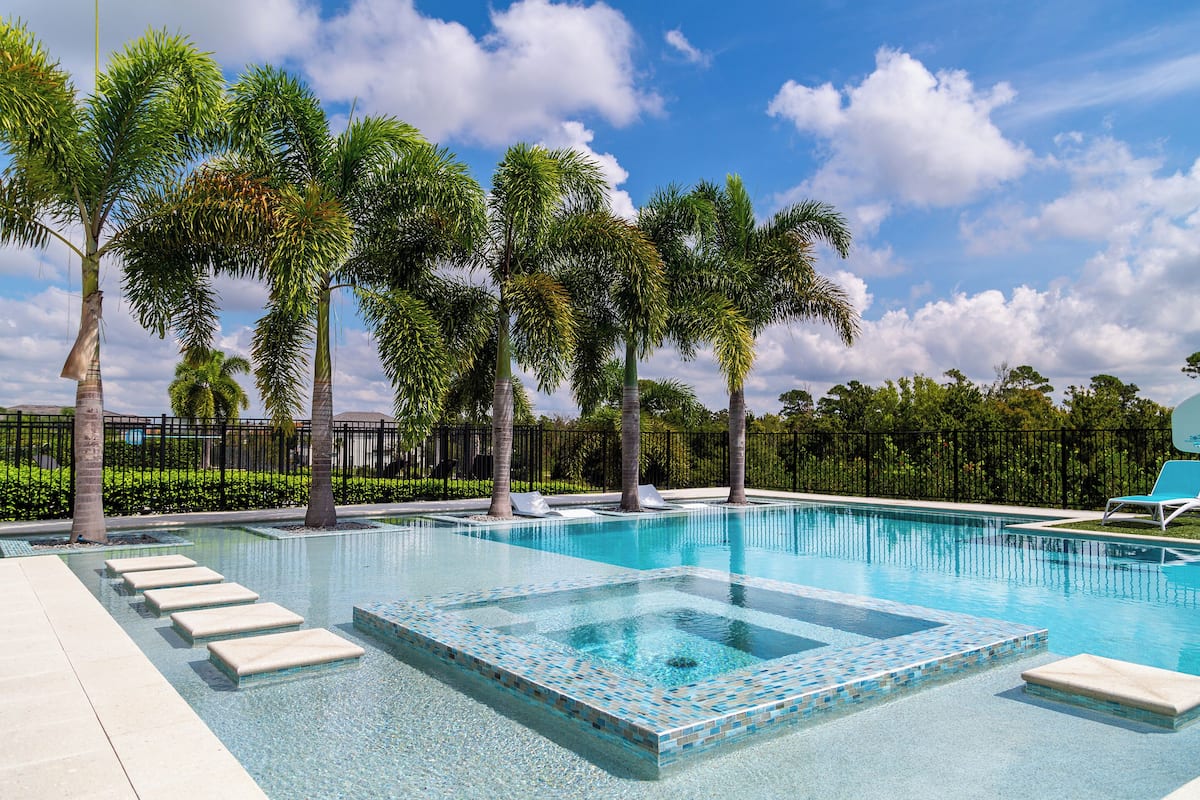 A swimming pool with clear blue water, surrounded by palm trees and a black fence.