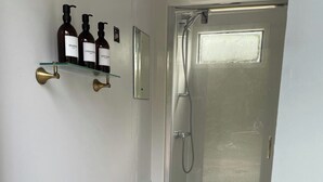 A bathroom with a glass shower door, a brass doorbell, and three bottles of shampoo on a glass shelf. - Trethowels Grey Hidden hut (Saint Austell)