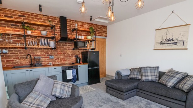 A compact living space with a kitchen area, grey sofas, and a hanging artwork. - Sea Spell Loft (Hornsea)