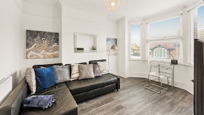 A modern living room with a sectional sofa, artwork on the walls, and a large window with blinds. - Modern 4BR Victorian Home near Luton Airport (Luton)