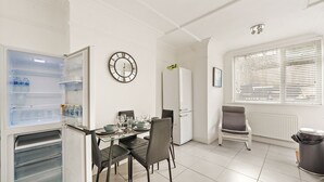 A compact kitchen with a refrigerator, a dining table with chairs, a clock, and a window with blinds. - Modern 4BR Victorian Home near Luton Airport (Luton)