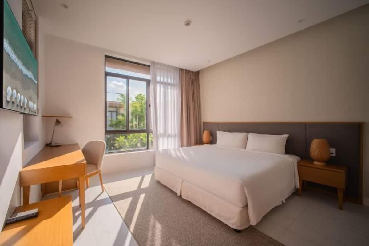 A modern hotel room with a large bed, a desk, a chair, a nightstand, and a window with curtains.