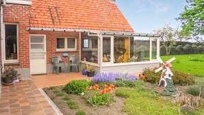 Una casa con fachada de ladrillo, techo de tejas y un invernadero con paredes de vidrio. Se observa un jardín con flores y un molino.