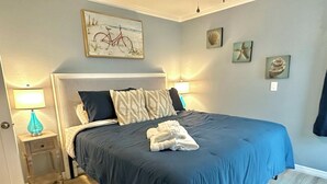 A neatly made bed with a blue bedspread, a headboard with a painting, and a small bedside table with a lamp.