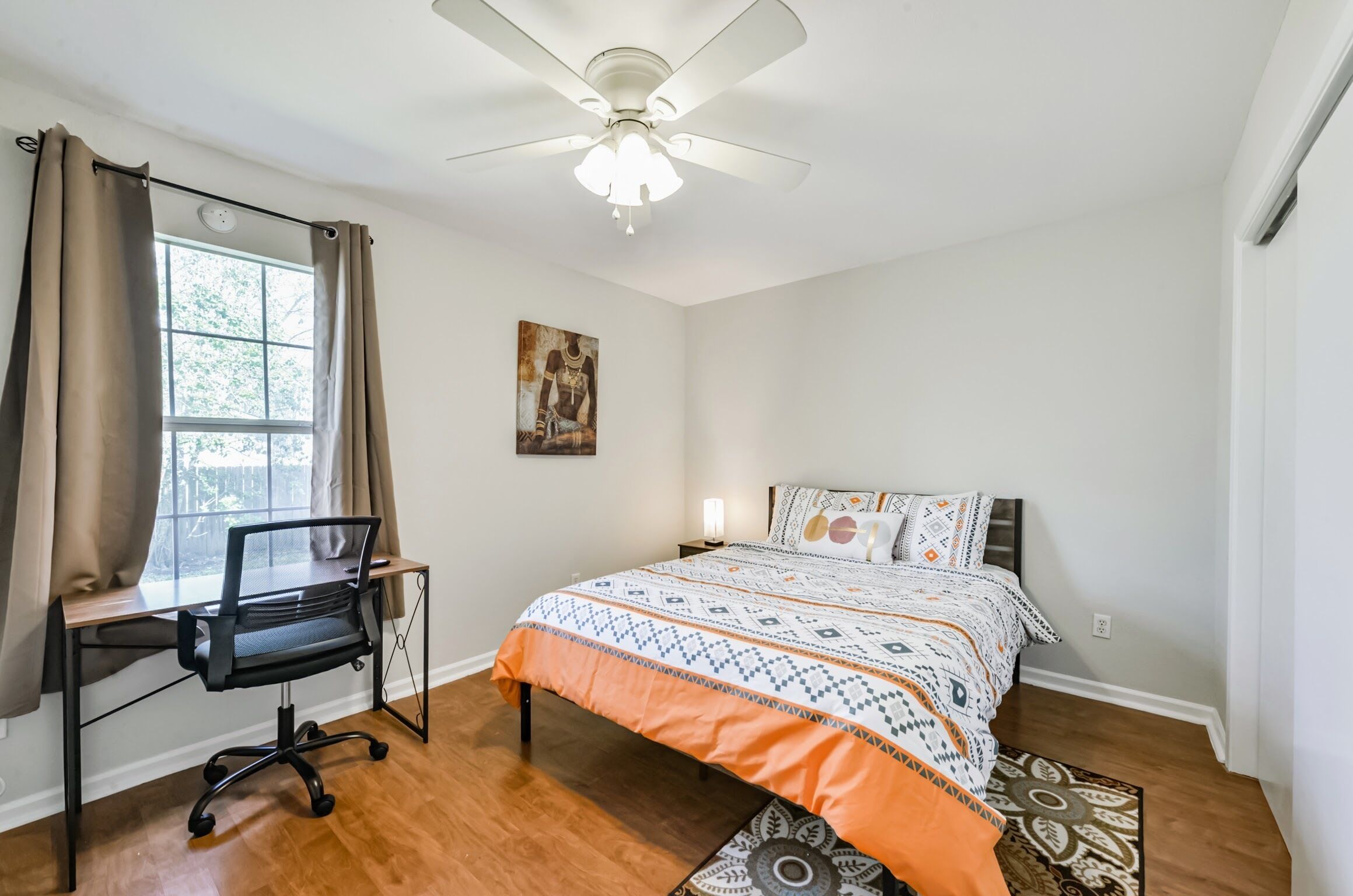 A bedroom with a bed, a desk with a chair, a ceiling fan, and a window with curtains.
