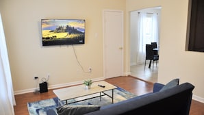 Una sala de estar con un televisor de pantalla plana montado en la pared, una mesa de centro, un sofá y un comedor al fondo.