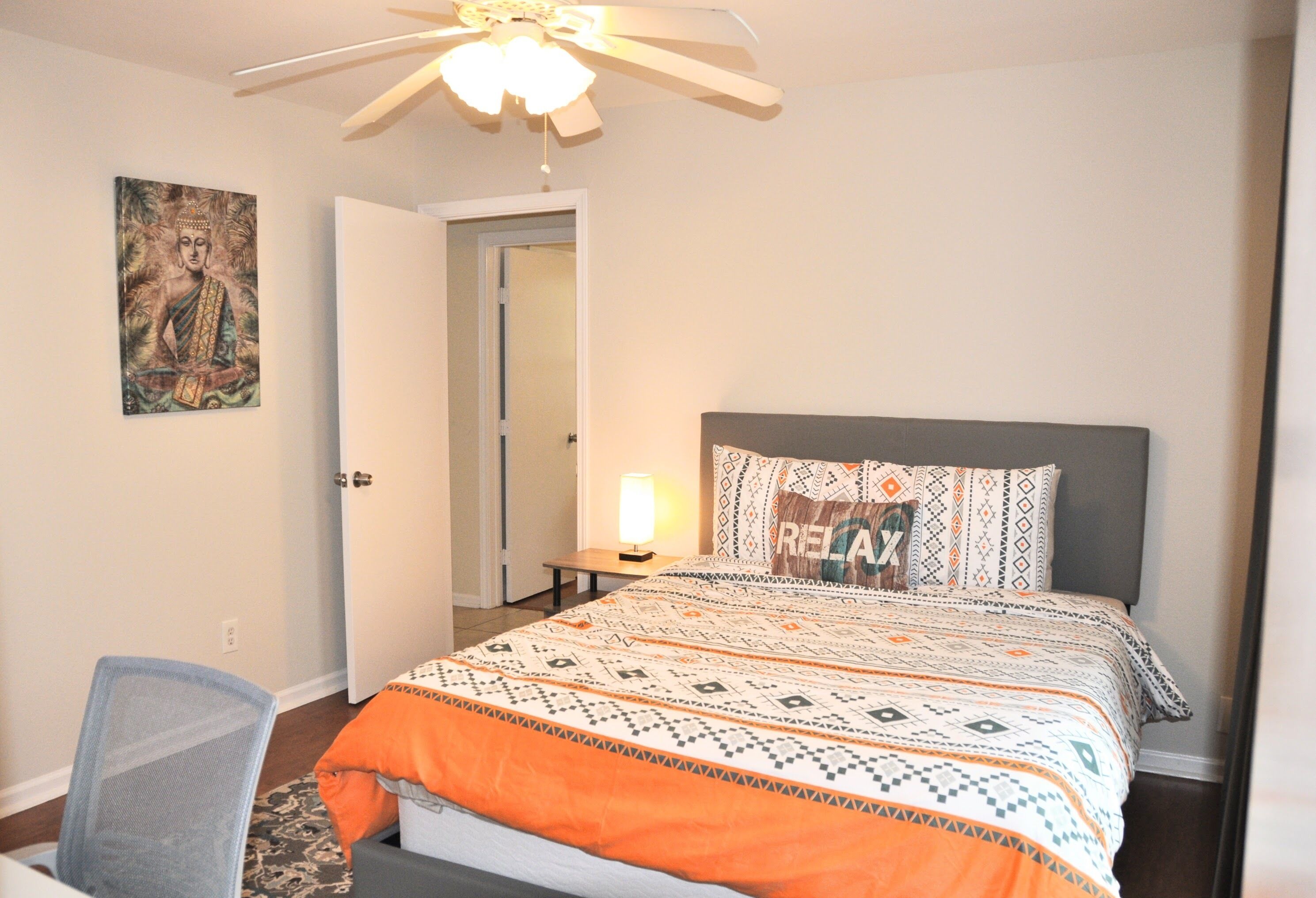 A bedroom with a bed, a desk, a chair, a ceiling fan, and a door.