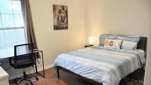 Un dormitorio con cama, un escritorio con silla, una mesita de noche con lámpara y un cuadro en la pared.