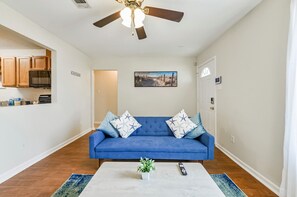 A living room with a blue sofa, a ceiling fan, a kitchen area, and a door with a sign.