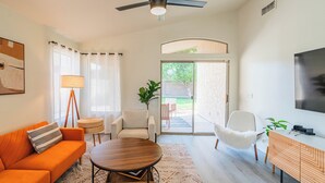 Uma sala de estar moderna com sofá, mesa de centro e cadeiras, um ventilador de teto, televisão e uma porta de vidro deslizante que dá para um pátio.