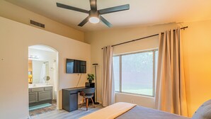 Quarto com cama, escrivaninha, cadeira, ventilador de teto, janela com cortinas e vista para o exterior.