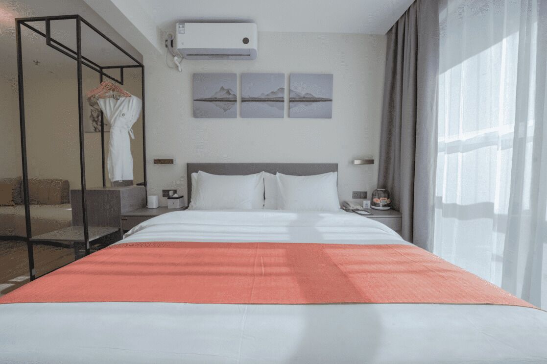 A hotel room with a large bed, a glass partition, a mounted air conditioner, and a view of mountains through the window.