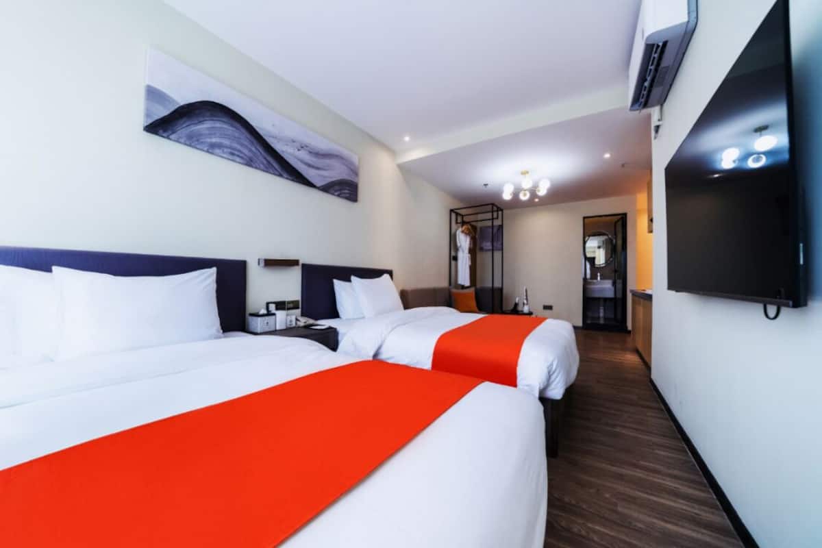 Habitación de hotel con dos camas, televisor, un cuadro en la pared y un pasillo que conduce al baño.