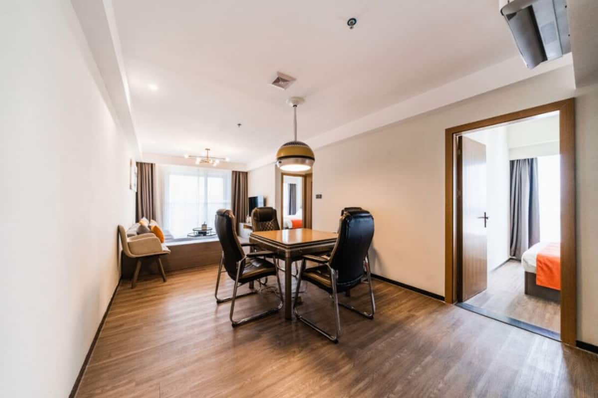 Habitación de hotel con piso de madera, una mesa de comedor con sillas, un espejo grande y una cama con manta naranja.