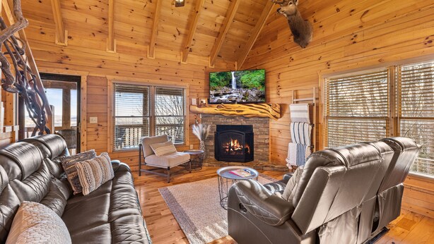 A log cabin interior with a fireplace, leather sofas, a television, and a deer head mounted on the wall. - Above & Beyond - Cabin w/ Massive Views Near Helen (Cleveland)