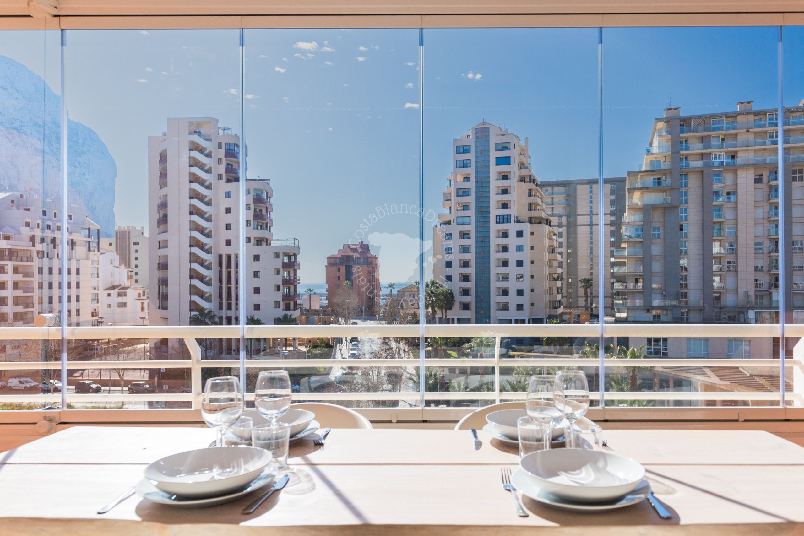Una mesa de comedor con platos, vasos y cubiertos, con vista a un paisaje urbano de edificios modernos.