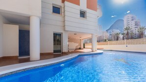 A swimming pool with a tiled edge, a white building with balconies, and a mountain in the background.