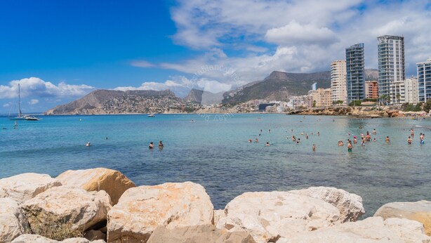 A coastal cityscape with clear blue waters, rocky shoreline, and modern buildings.