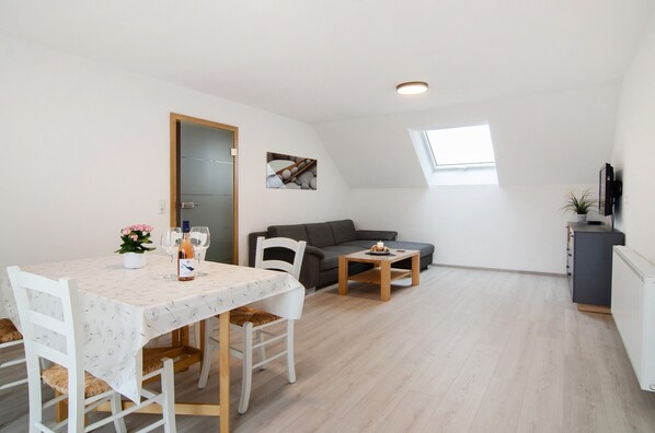 A modern living room with a dining area, a gray sofa, a small table, and a TV on a cabinet.