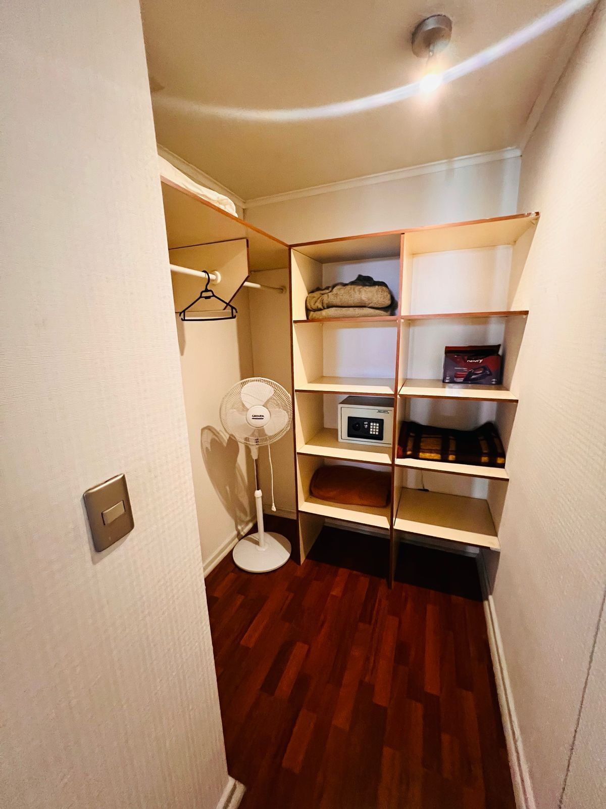 Un pasillo estrecho con suelo de madera, una pared blanca y un estante empotrado con diversos objetos.