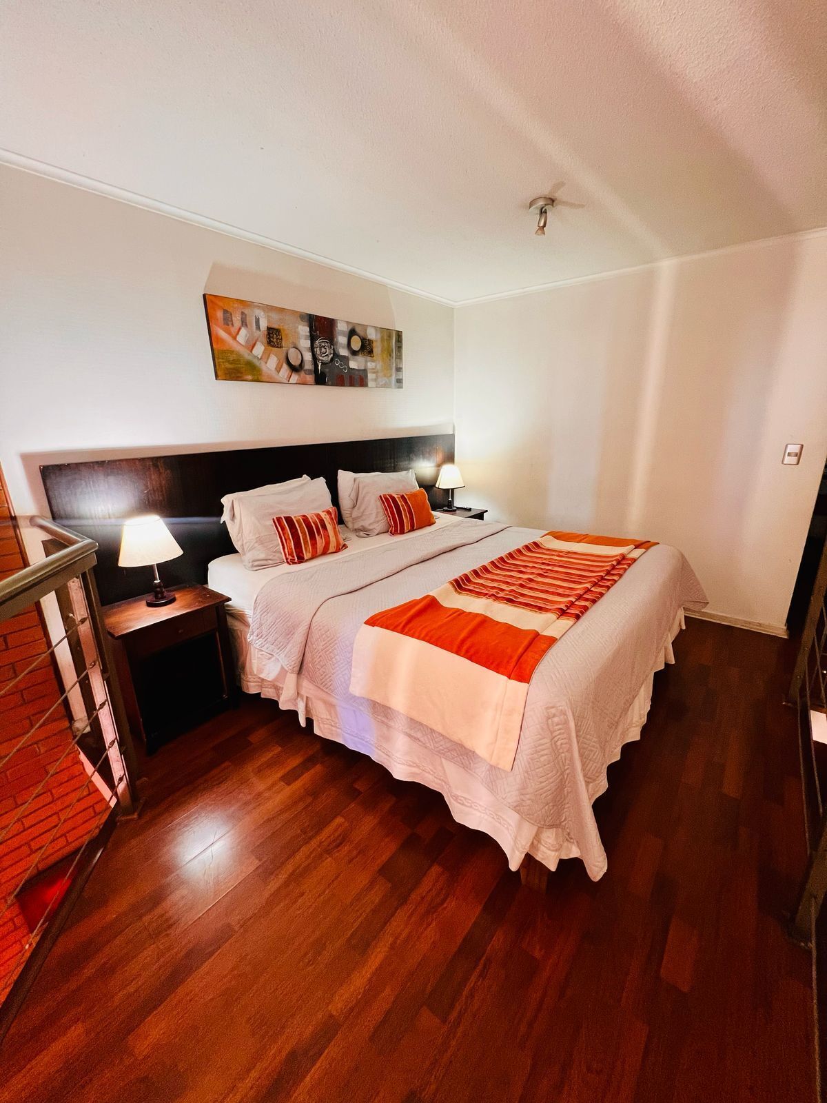 A bedroom with a large bed, a nightstand with a lamp, a headboard, and a painting on the wall.