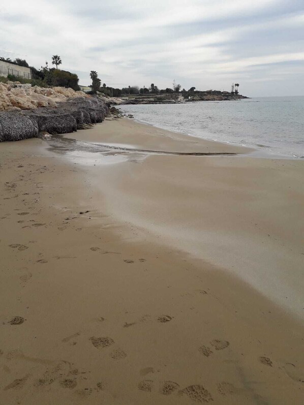 Une plage de sable parsemée d’empreintes, une digue rocheuse et une mer calme sous un ciel nuageux.