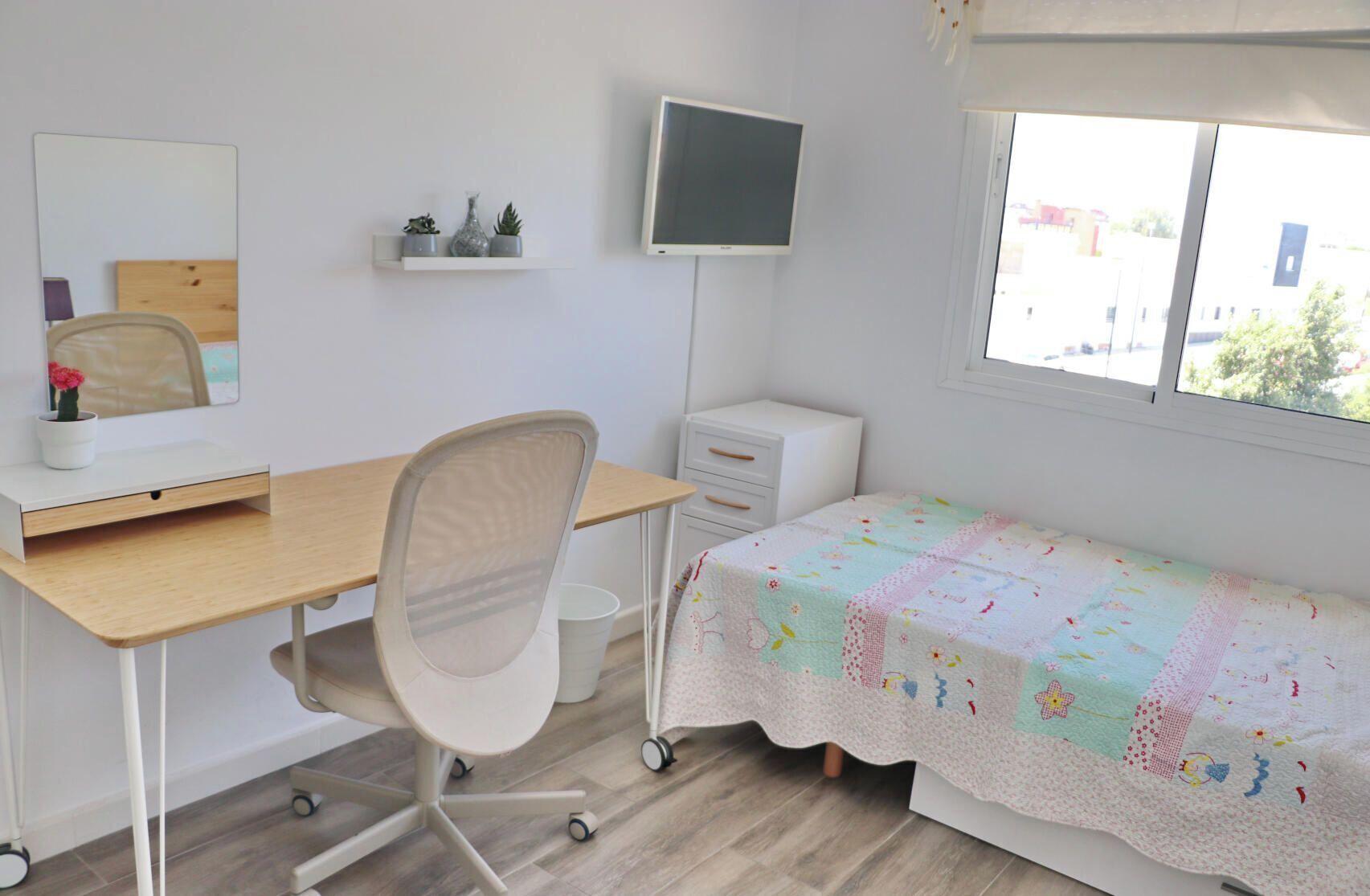 A bedroom with a desk, chair, bed, TV, and window.