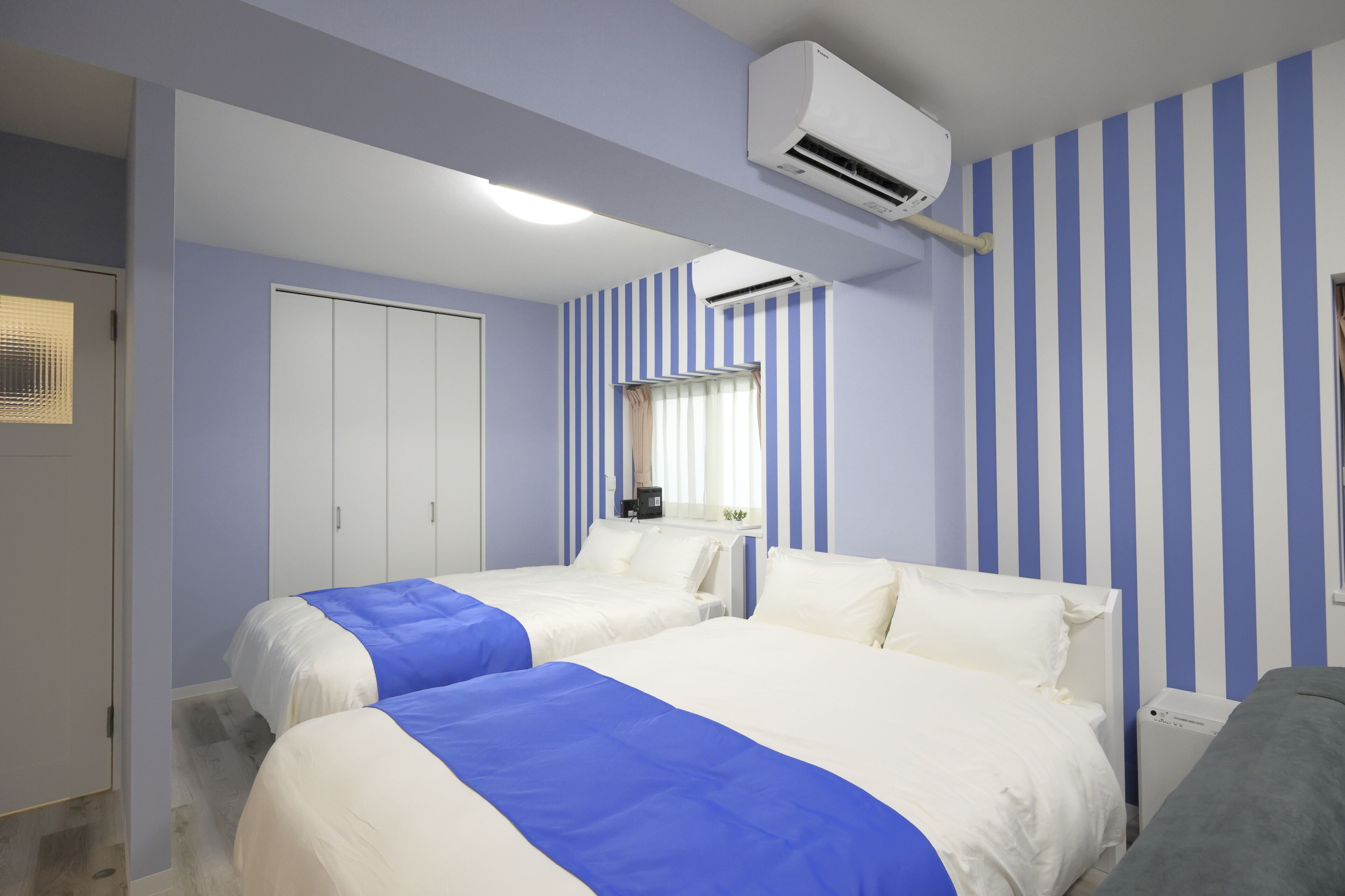 A hotel room with two beds, a wardrobe, an air conditioning unit, and a window with curtains.