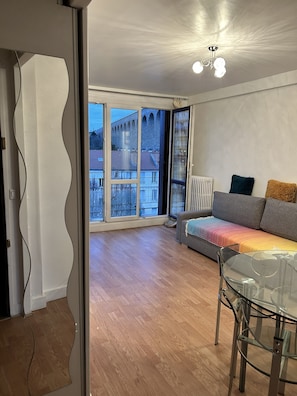 A spacious living room with a large window, a sofa, a glass table, and a radiator.