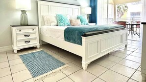 Un dormitorio con cama blanca, mesita de noche, alfombra azul y blanca estampada, y vista al balcón exterior.