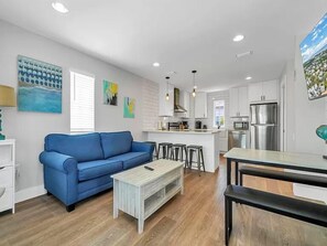 A modern living room with a blue sofa, a wooden coffee table, and a kitchen area with white cabinets and stainless steel appliances.
