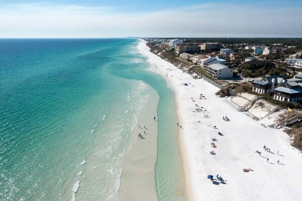 An aerial view of a beach with clear turquoise waters, a sandy shore, and buildings along the coastline.