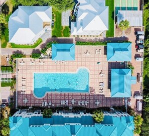 An aerial view of a resort with a central swimming pool, multiple buildings with blue roofs, and a parking area.