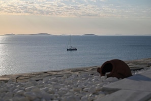 A calm sea with a sailboat in the distance, a rocky shoreline, and a clear sky. - Iliada Suites Adults Only (Naxos)