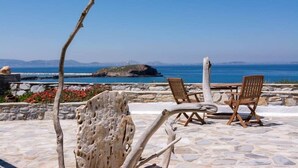 An outdoor seating area with stone flooring and wooden chairs, overlooking a coastal landscape with clear skies. - Iliada Suites Adults Only (Naxos)
