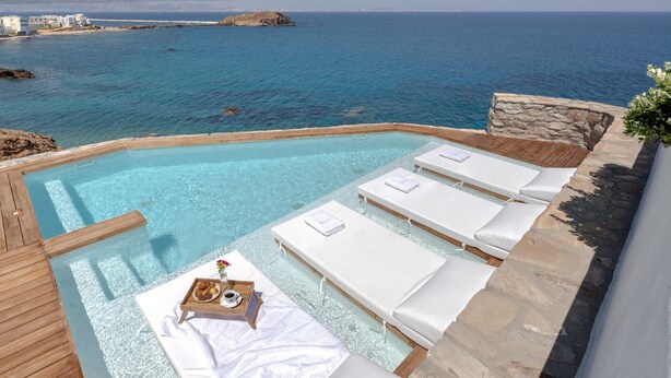 An infinity pool with white lounge chairs and a tray table with snacks. - Iliada Suites Adults Only (Naxos)